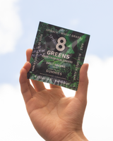 Hand holding a package of 8 Greens Daily Greens gummies against a clear sky.