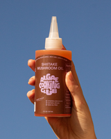 Hand holding a bottle of Algae Cooking Club's Shitake Mushroom Oil against a blue sky.