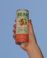 Hand holding a can of Bear Maple Farms Sparkling Ginseng Tonic in Ginger Citrus against a blue sky.