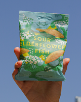 Hand holding a green package of Bon Bon's 'Sour Elderflower Fish' against a clear blue sky.