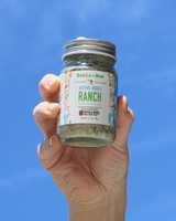 Hand holding a jar of Burlap and Barrel 'Ranch' seasoning against a blue sky