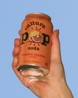 Hand holding a can of Culture Pop soda Orange Mango & Lime flavor, against a blue sky.
