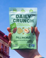 Hand holding a package of 'Daily Crunch' Dill Pickle Sprouted Nut snack against a blue sky.