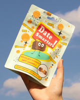 Hand holding a package of 'Date Smarter!' Chocolate Covered Date Caramels against a blue sky.