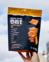 Package of Every Body Eat Cheese-less Thins held against a blurred cityscape background.
