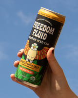 Hand holding a can of Freedom Fluid's Yaupon Iced Tea in Express Your Zest flavor against an open a sky.
