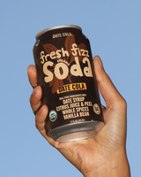 Hand holding a can of Fresh Fizz Organic Date Cola against a blue sky.