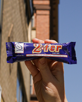 Hand holding a Go Max Go 2fer vegan candy bar against a blurred cityscape.