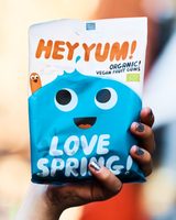 Hand holding a package of Hey Yum 'Love Spring' organic gummies against a blurred background.