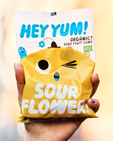 Hand holding a bag of Hey Yum! Sour Flower organic gummies against a blurred background.