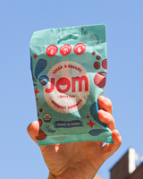 Hand holding a packet of Jom Gourmet Retro Cola Gummies against a clear blue sky.