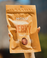 Hand holding a package of Moku Foods Sweet & Spicy Mushroom Jerky against a blurred background.