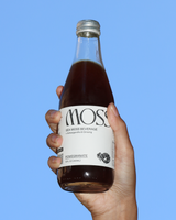 Hand holding a bottle of Moss Pomegranate Sea Moss drink against a blue sky.