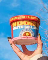 Hand holding a container of NOOISH Matzo Ball Soup against a blue sky.