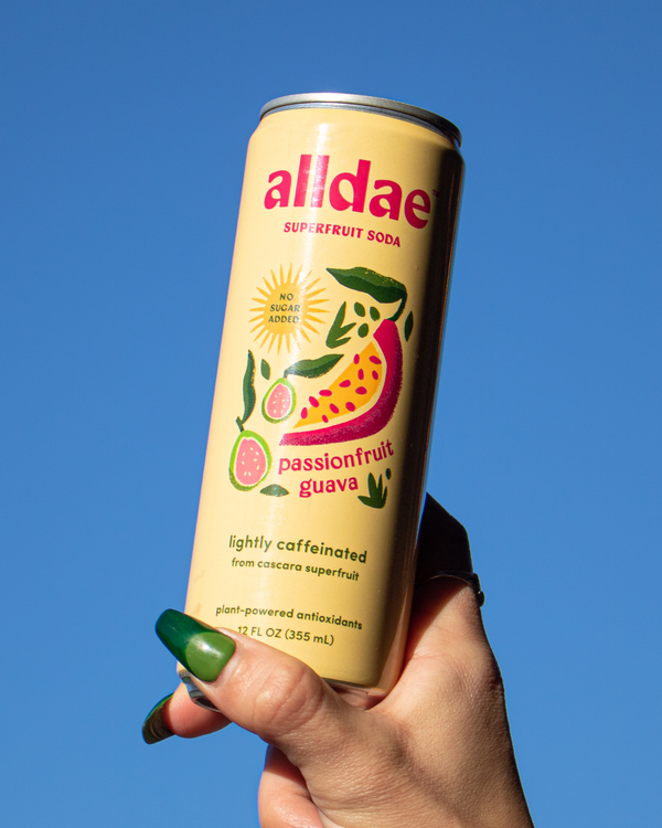 Hand holding a can of Alldae Passionfruit Guava Superfruit Soda against a blue sky.