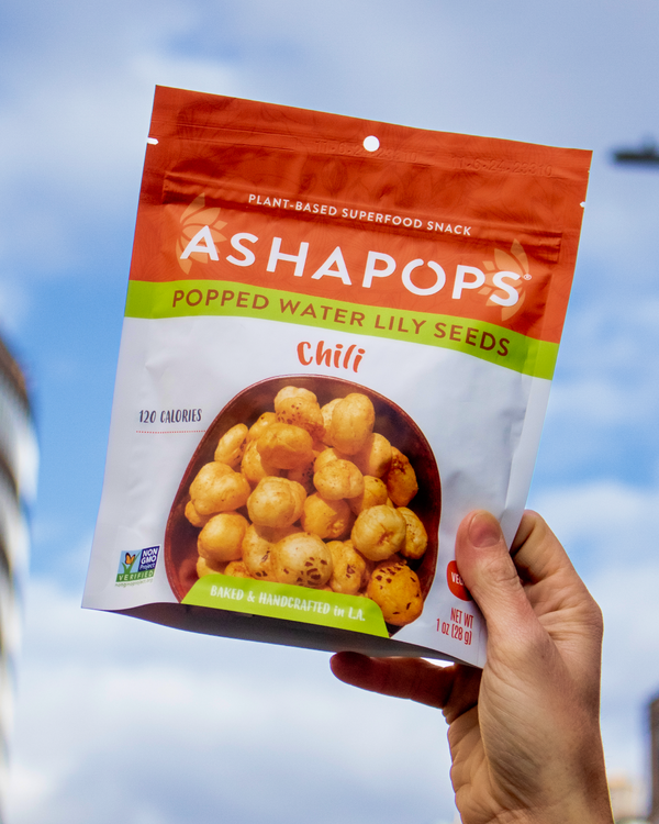 Hand holding a package of Ashapops Chili flavored popped water lily seeds against a blue sky.