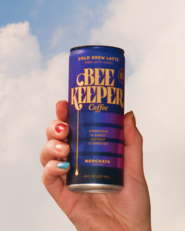 Hand holding a can of Bee Keeper Coffee in Horchata against a cloudy sky.