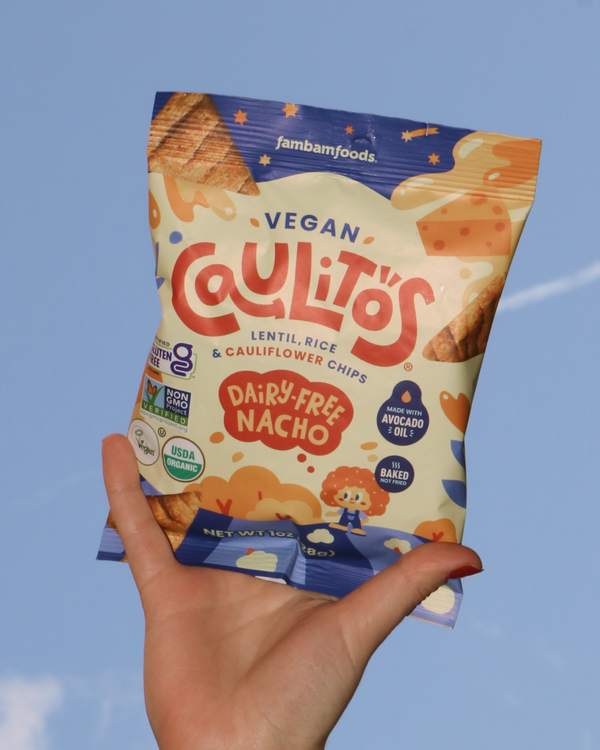 Hand holding a package of Vegan Caulitos Dairy-Free Nacho chips against a blue background.
