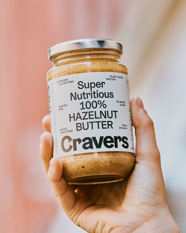 Hand holding a jar of Cravers 100% Hazelnut Butter with a blurred background.