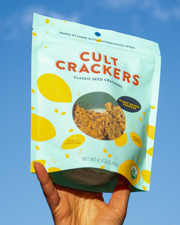 Hand holding a package of Cult Crackers Classic Seed Crackers against a blue sky background.