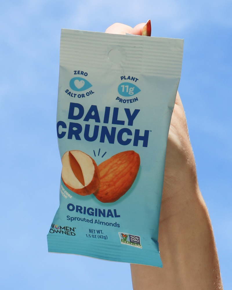 Hand holding a packet of Daily Crunch Original Sprouted Almonds against a blue sky.