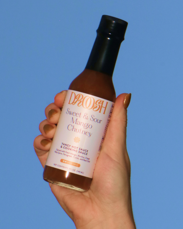 Hand holding a bottle of Droosh Sweet & Sour Mango Chutney against a blue sky.