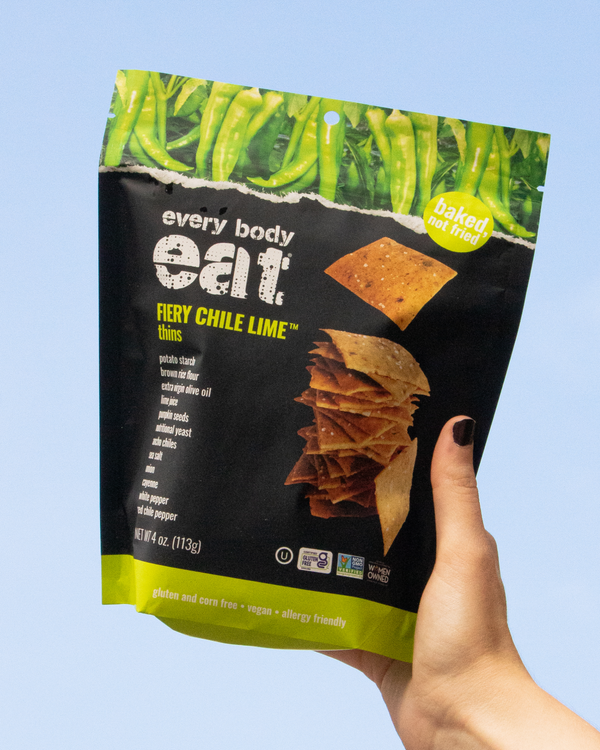 Hand holding a bag of Every Body Eat Fiery Chile Lime thins against a light blue sky.