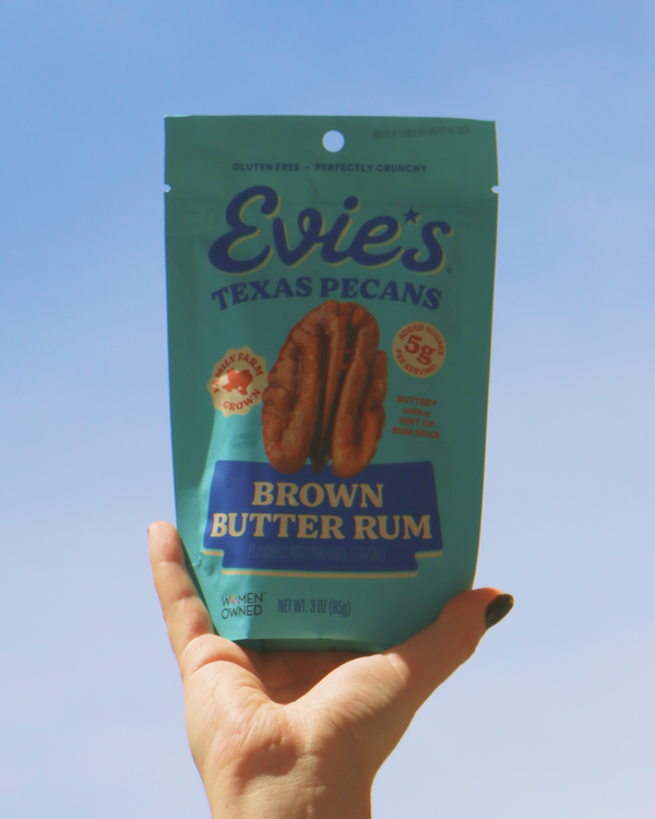 Hand holding a package of Evie's Texas Pecans Brown Butter Rum flavor against a clear blue sky.