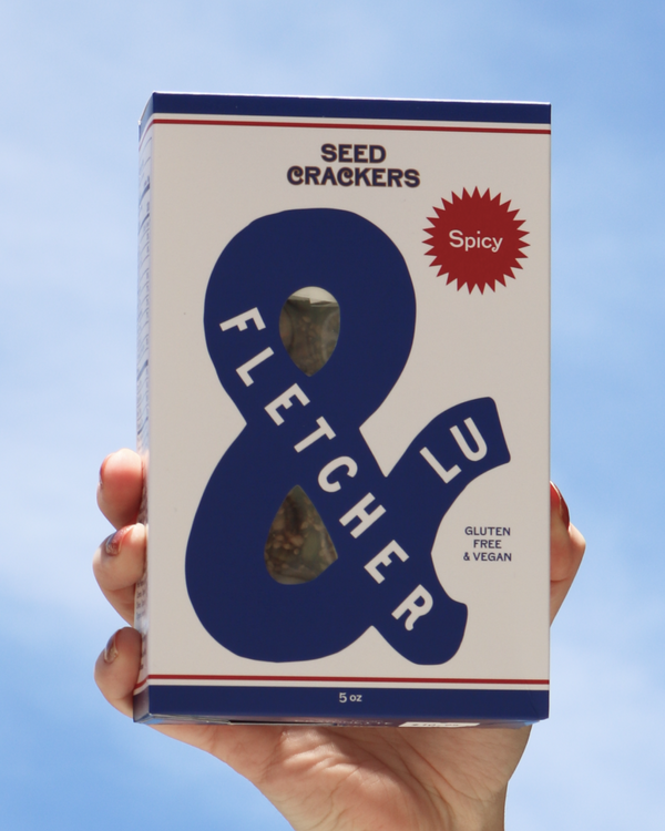 Hand holding a box of Fletcher & Lu Spicy Seed Crackers against a blue sky. 