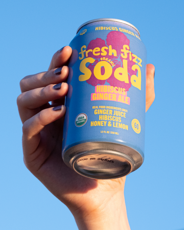 Hand holding a can of Fresh Fizz Soda in Hibiscus Ginger Ale against an open sky.