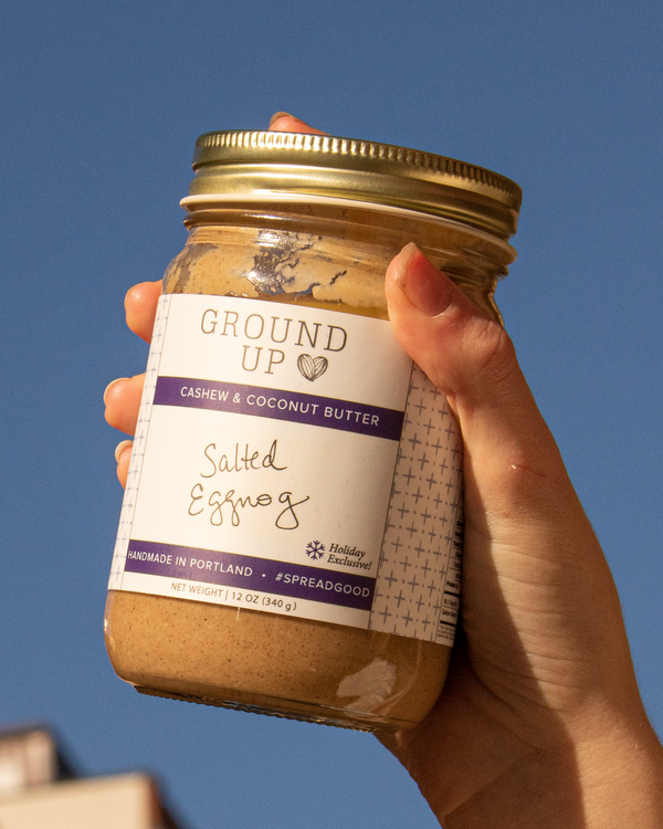 Hand holding a jar of Ground Up Salted Eggnog Cashew & Coconut butter against an open sky.