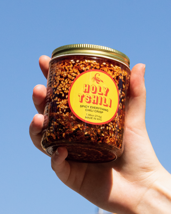 Hand holding a jar of Holy Tshili Spicy Everything Chili Crisp against an open sky.
