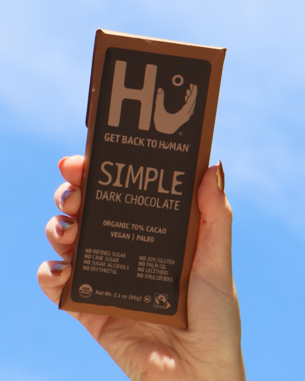 Hand holding a bar of HU Simple Dark Chocolate against a clear blue sky