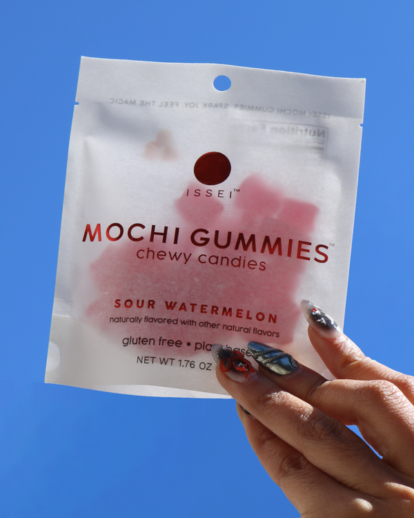 Hand holding a package of Issei Mochi Gummies Sour Watermelon against a blue sky.