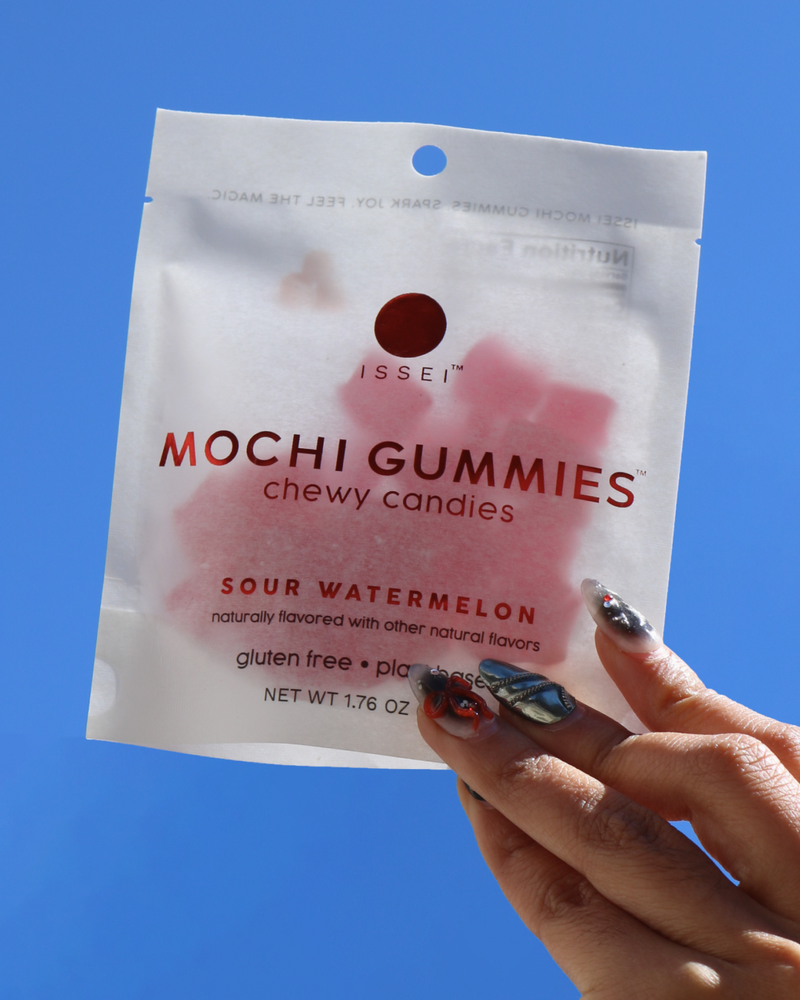 Hand holding a package of Issei Mochi Gummies Sour Watermelon against a blue sky.