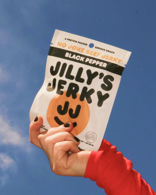 Hand holding a package of Jilly's Black Pepper Jerky against a blue sky.