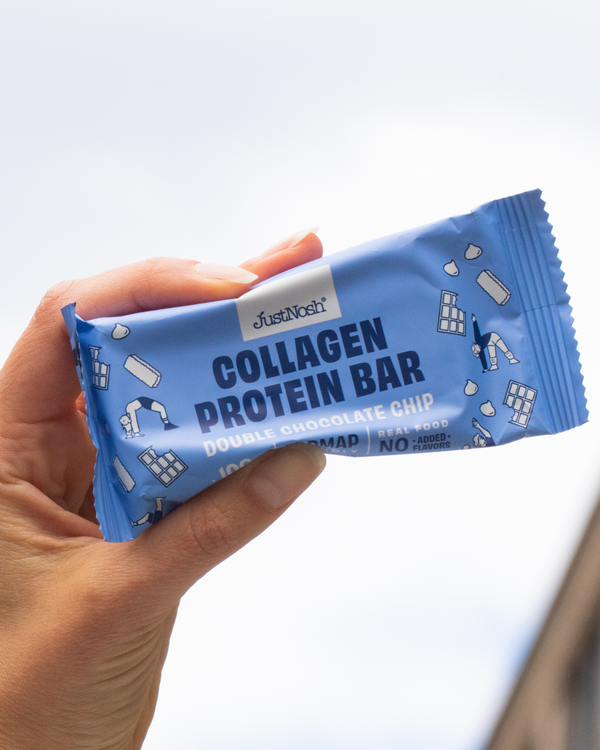 Hand holding a JustNosh Double Chocolate Chip Collagen Protein Bar against a blurred background. 