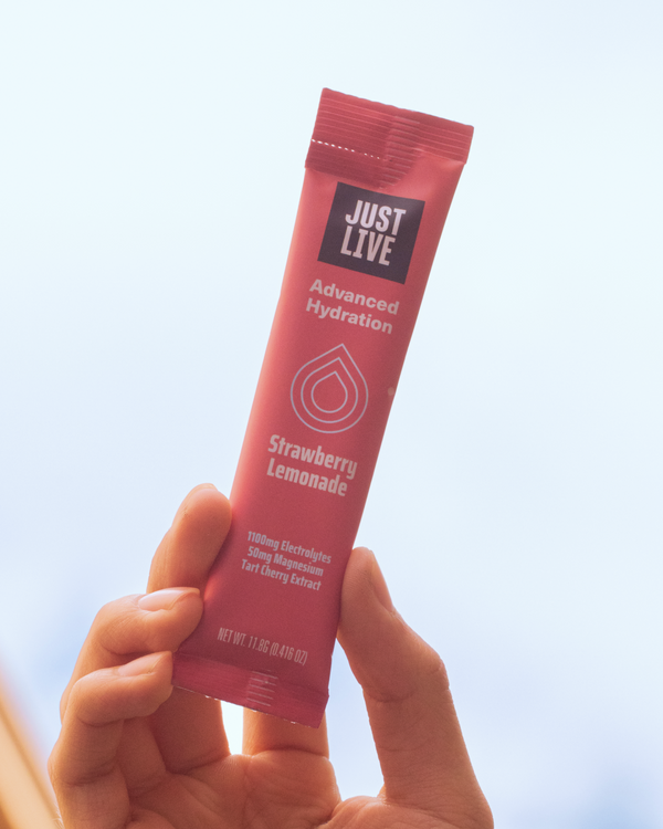 Hand holding a package of Just Live Advanced Hydration in Strawberry Lemonade against a grey sky. 