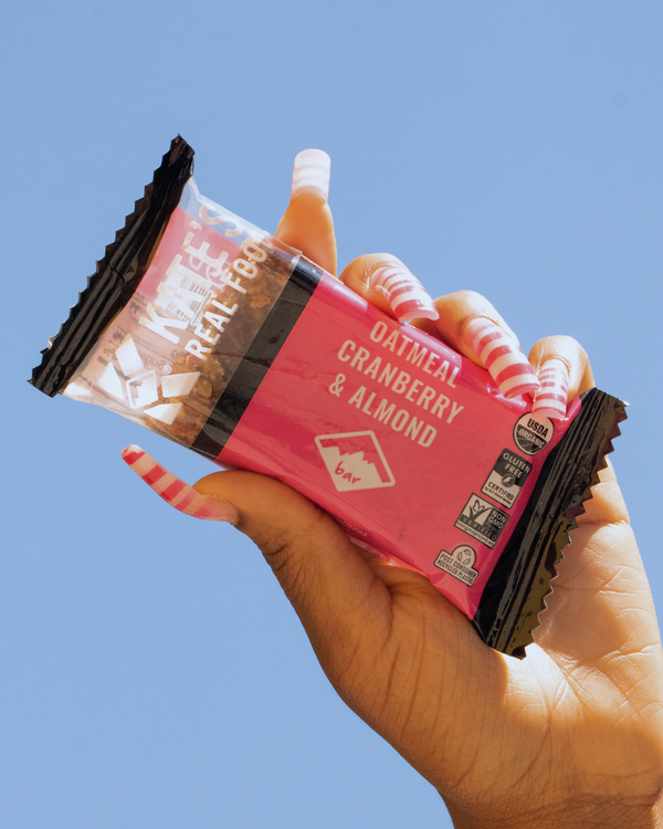 Hand holding a bar of Kate's Real Food Oatmeal Cranberry & Almond granola bar against an open sky.