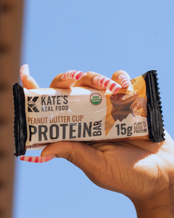 Hand holding a Kate's Real Food Peanut Butter Cup Protein Bar against a blue sky background