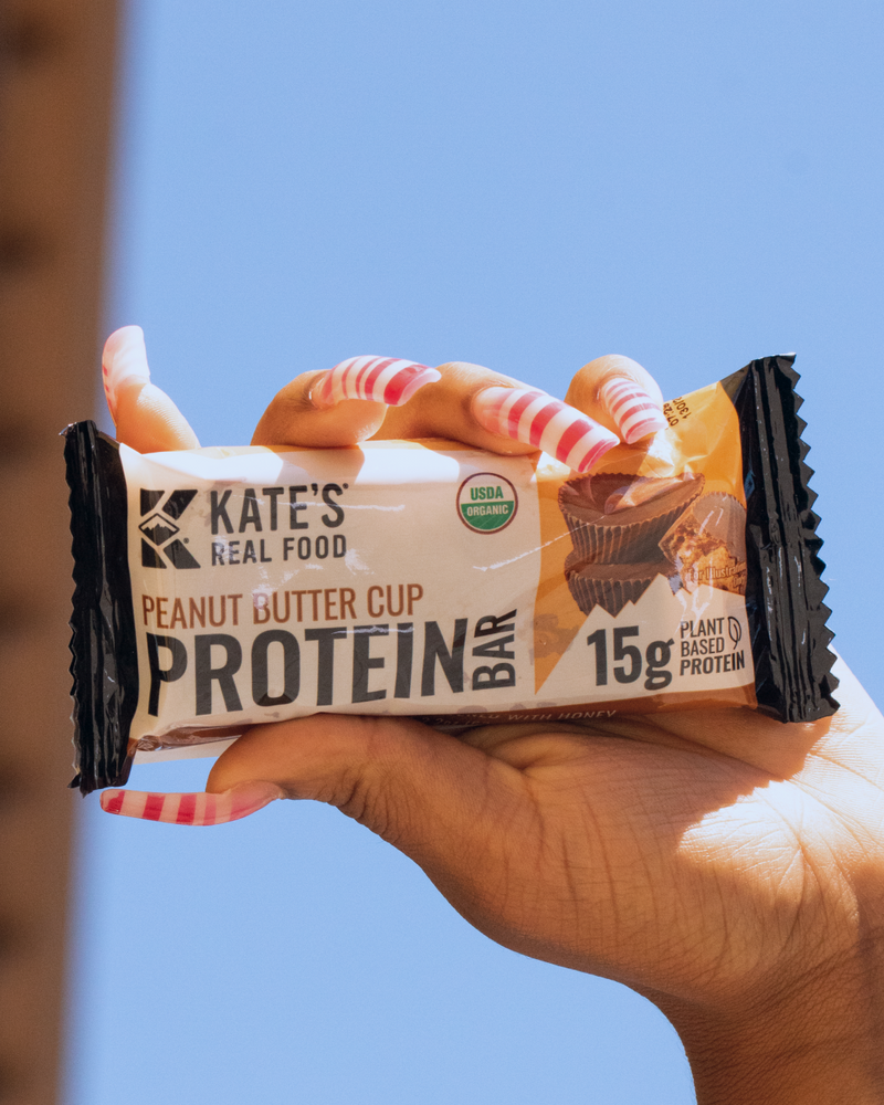 Hand holding a Kate's Real Food Peanut Butter Cup Protein Bar against a blue sky background