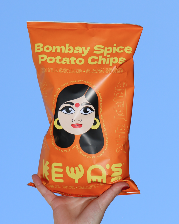 Hand holding a bag of Keya's Bombay Spice potato chips against an open sky.
