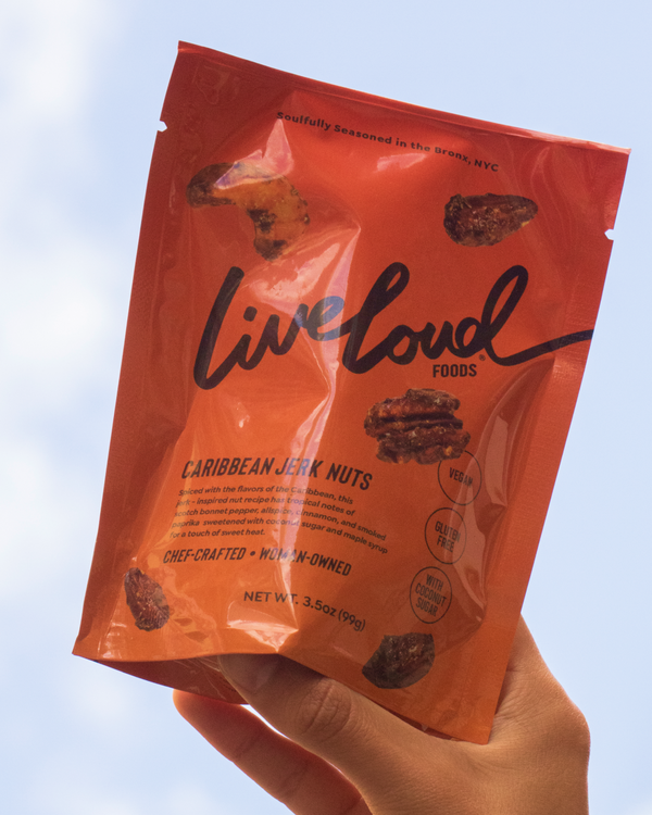 Hand holding a bag of Live Loud Foods Caribbean Jerk Nuts against an open sky.