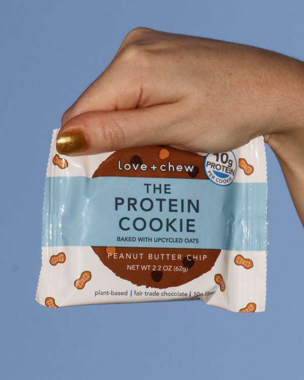 Hand holding a Love + Chew Peanut Butter Chip Protein Cookie against a blue sky.