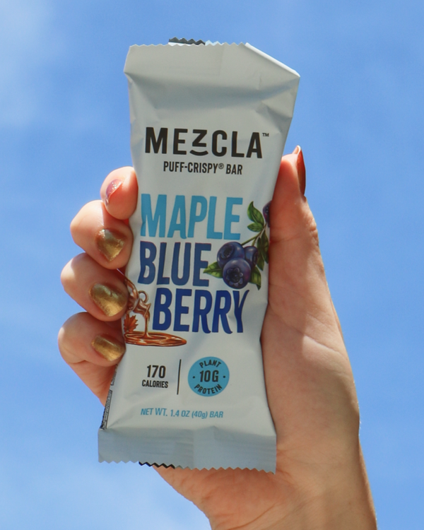 Hand holding a Mezcla Maple Blueberry snack bar against a clear blue sky.