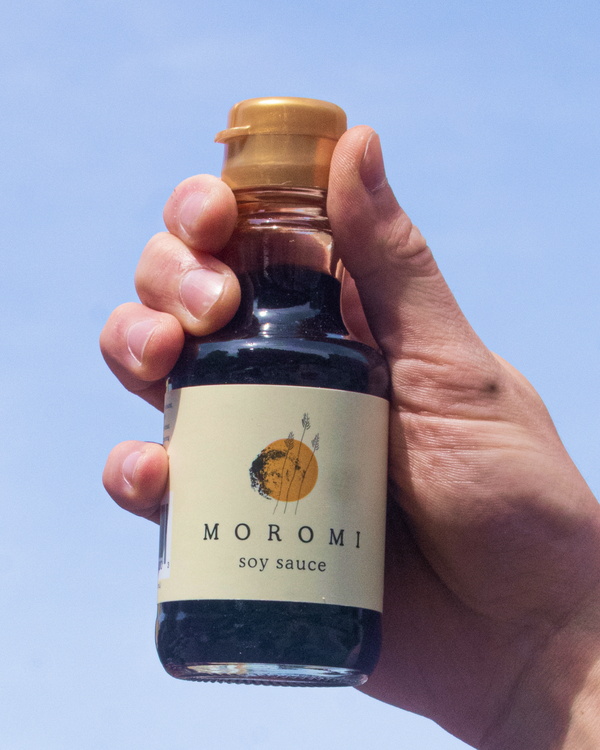 Hand holding a bottle of Moromi Shoyu Soy Sauce against a blue sky.